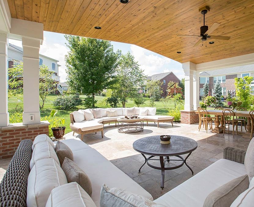 Covered patio with wooden ceiling, featuring L-shaped white cushioned sofas, circular coffee tables, and a dining set. Overlooks a sunny, green backyard.